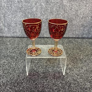 May include: Two matching ruby red glass goblets featuring delicate gold filigree scrollwork patterns on the bowls and circular bases. Each glass has a slender, textured stem and a gold-rimmed edge, presenting a consistent design across both pieces.