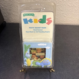 May include: A blue and yellow dinosaur photo magnet frame with the text "HOLSON kids". The frame has a hand-painted design and an easel back for self-standing display. The frame is packaged in a clear plastic case.