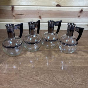 May include: Four clear glass pitchers with silver trim and black handles. Each pitcher has a round body and a silver-colored top. The handles are black and curved. The pitchers are arranged in a row on a wooden surface.