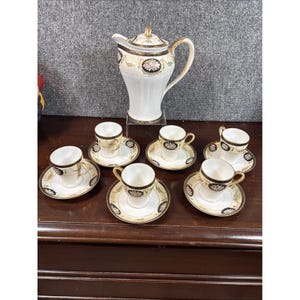 Może przedstawiać: Porcelanowy zestaw do kawy składający się z wysokiego dzbanka z pokrywką oraz sześciu pasujących filiżanek ze spodkami. Każdy element jest kremowy z czarnymi obwódkami, złotymi zdobieniami i motywami kwiatowymi. Zestaw ma spójny wygląd.