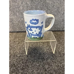 Puede incluir: Una taza de cerámica con un diseño de vaca azul y blanco. La taza tiene un interior azul claro y un asa blanca. La vaca está representada con contornos azules y manchas blancas. La base de la taza es verde.