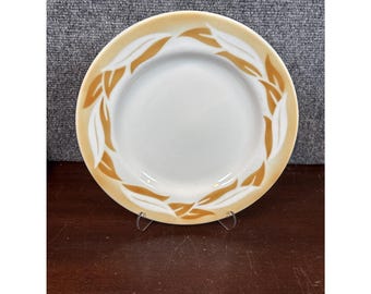 Puede incluir: Un plato de cerámica blanco con un borde naranja claro. El borde está decorado con un patrón repetido de hojas estilizadas de color naranja claro. El plato se exhibe sobre un soporte metálico.