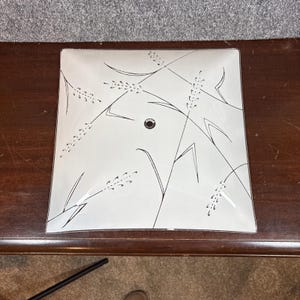 May include: Square, frosted glass ceiling light fixture with a black and white wheat stalk design. The fixture has a small, round, black center detail. The light is sitting on a dark brown wooden surface.