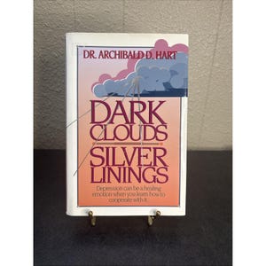 May include: A hardback book titled Dark Clouds Silver Linings by Dr. Archibald D. Hart. The cover features a gradient background from white to orange with a graphic of blue and pink clouds. The text reads: Depression can be a healing emotion.