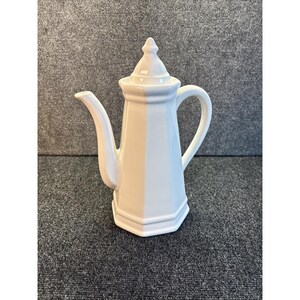 May include: A white ceramic coffee pot with a long, curved spout and a handle. The pot has a geometric design with a pointed lid. The pot is likely used for serving coffee or other hot beverages.