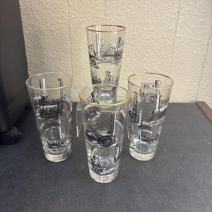 May include: Four clear drinking glasses with gold rims, each featuring a black and white steam engine illustration. The glasses are different heights and shapes, ideal for water or other drinks.