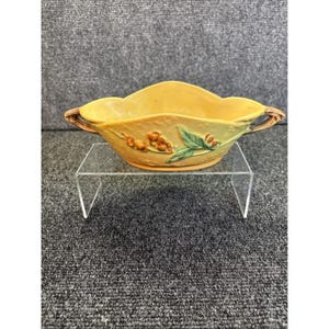 Puede incluir: Una fuente de cerámica amarilla con borde festoneado y dos asas. El plato está decorado con un diseño en relieve de bayas y hojas. Las asas tienen un aspecto marrón, similar a la madera. El plato se exhibe sobre un soporte de acrílico transparente.