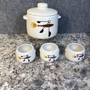 May include: A cream-colored ceramic kitchen set featuring a lidded pot and three small bowls. Each piece is decorated with a brown, yellow, and black utensil design. The set has a speckled finish and a vintage aesthetic.