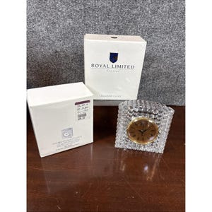 May include: A crystal crescent clock with a gold-tone face and a textured, square crystal base. The clock is displayed next to two white boxes, one labeled "ROYAL LIMITED Crystal" and the other with "SQUARE CRESCENT CLOCK".