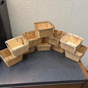 May include: A collection of ten small, square wooden baskets. The baskets are made of light-colored wood and have a simple, open design. They are arranged in a slightly curved pattern on a dark surface, suggesting they are for display or storage.