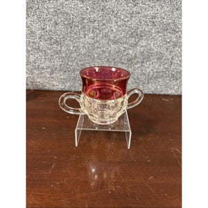 Puede incluir: Una pequeña taza de cristal transparente con dos asas. El borde superior es de color rojo intenso con un acento dorado. La taza está sobre un soporte de plástico transparente. La taza tiene un patrón decorativo en la mitad inferior.