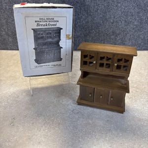May include: A miniature wooden breakfront doll house furniture piece. The breakfront is brown with three upper cabinets with window-like designs and three lower cabinet doors. The box next to it reads "DOLL HOUSE MINIATURE WOODEN Breakfront".