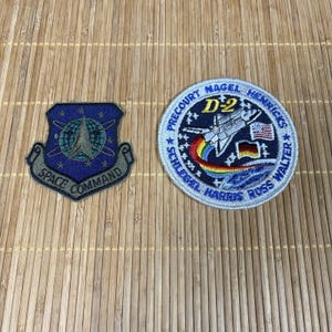 以下が含まれることがあります： 2つの刺繍パッチ。1つは、銀と緑のデザインと「SPACE COMMAND」の文字が入った青いシールドです。もう1つは、スペースシャトル、虹、「D2」、「PRECOURT」、「MAGEL」、「HENRICKS」、「SCHLEGEL」、「HARRIS」、「ROSS WALTER」の文字が入った円形のパッチです。