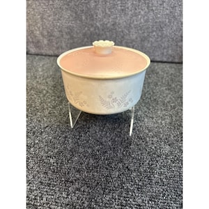 May include: A vintage, off-white casserole dish with a pink lid and floral design. The dish sits on a clear, angled stand. The lid has a flower-shaped handle. The dish has a delicate, vintage aesthetic.