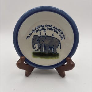 May include: A round ceramic plate with a blue rim featuring a hand-painted illustration of two gray elephants standing on a patch of green grass. Above the elephants, the text reads, Two of every sort shall thou bring into the Ark.