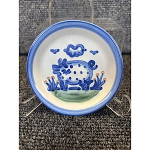 Puede incluir: Plato pequeño y redondo de cerámica con un diseño de cerdo pintado a mano. El plato tiene un centro blanco con un borde azul y una base verde. El cerdo es azul con lunares blancos, y el diseño incluye nubes y hierba azules.