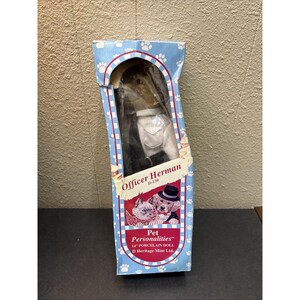 May include: A boxed "Officer Herman" porcelain doll from the "Pet Personalities" collection. The box is blue with paw prints and features illustrations of a cat and dog. The doll is wearing a white shirt and black tie. The box reads "14" PORCELAIN DOLL".