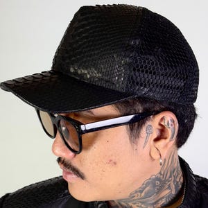 May include: A black leather baseball cap with a snakeskin pattern on the front and a mesh back. The cap is worn by a person with a tattoo on their neck.