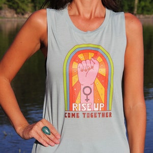 Può includere: Una canotta blu chiaro con una grafica arcobaleno di un pugno alzato e il testo "Rise Up Come Together".