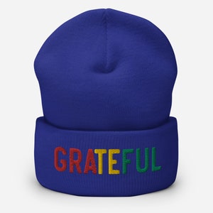 May include: A royal blue beanie with a fold-over cuff. The cuff has the word "GRATEFUL" embroidered in red, yellow, and green.