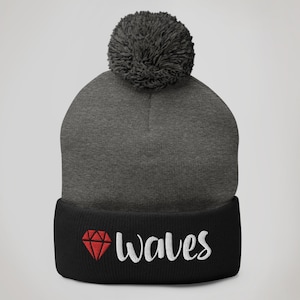 May include: Gray and black knit beanie with a pom pom on top. The beanie has a black cuff with a red diamond and the word "Wales" in white.