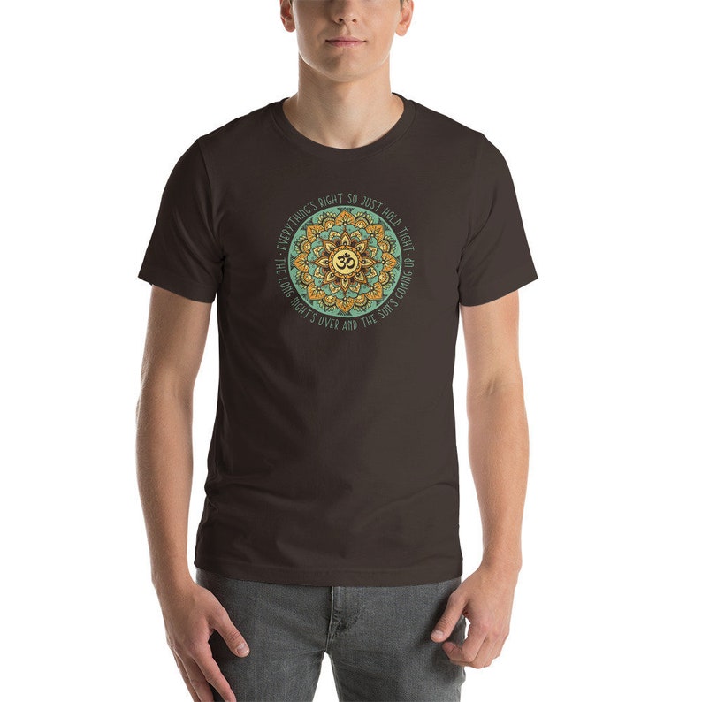 May include: A brown t-shirt with a green and gold mandala design featuring the Om symbol and the text "Everything's right so just hold tight - the long night's over and the sun's coming up."