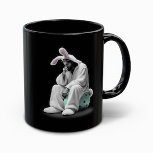 May include: Black ceramic mug featuring a black and white image of a person in a bunny costume. The person is sitting on a blue and green polka-dotted egg. The mug has a black handle.