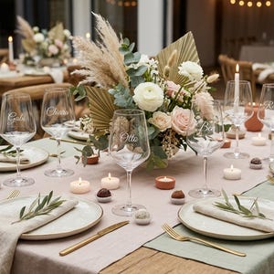 Könnte beinhalten: Ein festlich gedeckter Tisch mit Weingläsern, Tellern und Kerzen. Der Mittelpunkt ist ein Blumengesteck mit Rosen und Pampasgras. Jedes Weinglas ist mit einem Namen personalisiert. Die Tischdekoration ist elegant und für einen besonderen Anlass.