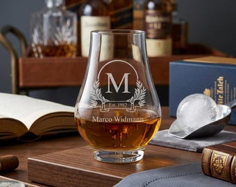 May include: A clear whiskey glass with a personalized design, filled with amber liquid. The glass features the letter "M" inside a laurel wreath, with the text "Est. 1982" and "Marco Widmann". The glass sits on a wooden surface.