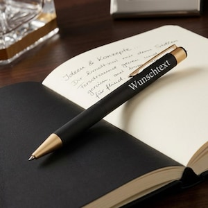 Könnte beinhalten: Ein mattschwarzer Kugelschreiber mit goldfarbenen Akzenten, einer strukturierten Griffzone und einem goldenen Clip. Auf dem Schaft ist der weiße Schriftzug "Wunschtext" aufgedruckt. Der Stift liegt diagonal auf einem offenen Notizbuch mit handschriftlichen Notizen.