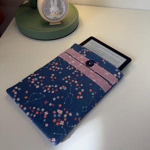 May include: A navy blue e-reader sleeve with a floral and constellation pattern. The sleeve has a pink band across the top and a dark blue button closure. The e-reader is partially visible inside the sleeve.