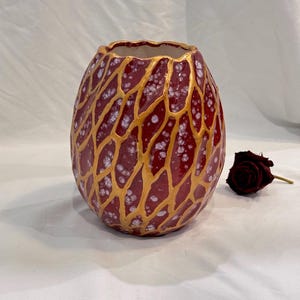 May include: A maroon ceramic vase with a gold webbed design and white speckles. The vase has a unique, textured surface and a ruffled rim. A single dark red rose rests beside the vase.