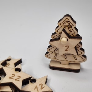 Könnte beinhalten: Adventskalender aus Holz in Form eines Weihnachtsbaums mit einem sternförmigen Ausschnitt, der die Zahl 2 anzeigt. Mehrere sternförmige Ornamente aus Holz mit Zahlen sind in der Nähe verstreut.