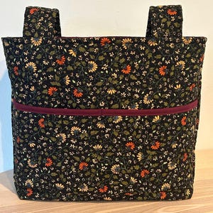 May include: A black tote bag with a floral pattern featuring orange, yellow, and white flowers. The bag has a burgundy pocket and matching handles. The bag is made of a quilted fabric.