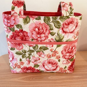 May include: A quilted fabric tote bag featuring a floral pattern with large pink and red roses, green leaves, and small orange blossoms on a cream background. The bag has two top handles and a horizontal coral-colored trim across the front pocket.