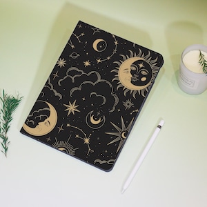 Pode incluir: Capa de tablet preta com um design celestial com um rosto de sol e lua, estrelas e constelações douradas. Uma caneta stylus branca e uma vela acesa também são visíveis.