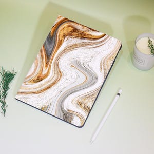 Op de afbeelding: Een tablet hoesje met een wervelend marmerontwerp in tinten wit, goud en grijs. Een witte stylus en een brandende kaars in een grijze houder zijn ook aanwezig. De hoes is rechthoekig.