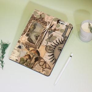 May include: A decorative tablet case featuring a collage of vintage-style illustrations. The design includes a framed landscape, a sun with an eye, butterflies, mushrooms, and a star. A white stylus and a candle are also visible.