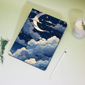 Pode incluir: Uma capa para tablet com um design de céu noturno, apresentando uma lua crescente, estrelas e nuvens em tons de azul e branco. Uma caneta stylus branca e uma vela com alecrim também estão presentes.