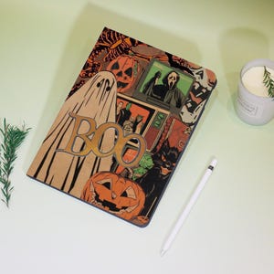 Può includere: Un taccuino a tema Halloween con una copertina marrone con un fantasma, zucche e la parola "BOO". Il taccuino presenta anche immagini di un gatto nero e un personaggio del film "Scream". Nello scatto sono presenti anche uno stilo bianco e una candela.