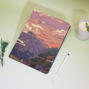 Könnte beinhalten: Eine Tablet-Hülle mit einem Landschaftsdesign mit Bergkette, Sonnenuntergang und Mond. Die Hülle ist in den Farbtönen Lila, Orange und Blau gehalten. Ein weißer Stift und eine Kerze sind ebenfalls sichtbar.