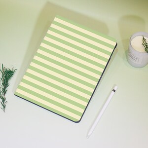 May include: A green and cream striped notebook lies on a light green surface, next to a white stylus and a lit candle in a white jar. A sprig of rosemary is also present. The notebook's cover features horizontal stripes.