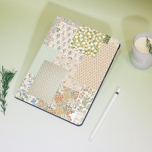 Könnte beinhalten: Eine Tablet-Hülle mit einem Patchwork-Design mit Blumenmustern in Grün, Gelb und Orange. Die Hülle hat einen dunkelblauen Rand. Ein weißer Stift und eine brennende Kerze mit Rosmarin sind ebenfalls zu sehen.