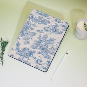 Può includere: Un taccuino rettangolare con un motivo toile de Jouy blu e bianco raffigurante scene pastorali. Il taccuino ha un bordo nero ed è accanto a uno stilo bianco e una candela accesa in un contenitore grigio con un rametto di rosmarino.
