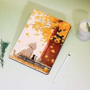 Op de afbeelding: Een notitieboek met een kattenillustratie. De omslag toont een grote witte kat en een kleine zwarte kat die op een stenen richel zitten, met een achtergrond van een deur en gele herfstbladeren. Een witte stylus en een kaars zijn ook zichtbaar.