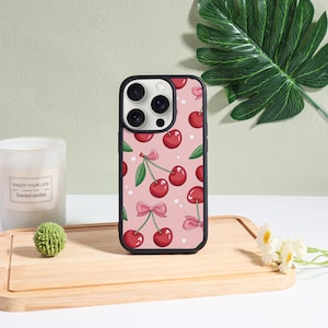 Op de afbeelding: Zwarte telefoonhoes met een roze achtergrond met een patroon van rode kersen, groene bladeren en roze strikken. De hoes heeft een zwarte rand en een camera-uitsparing. Een witte kaars en een kleine groene decoratieve bal staan op de achtergrond.