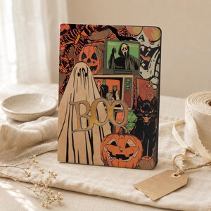 May include: A rectangular notebook featuring a Halloween-themed graphic print with a large ghost, orange jack-o'-lanterns, a black cat, and vintage television sets displaying horror imagery. The word BOO is printed in large, bold, gold-toned letters.