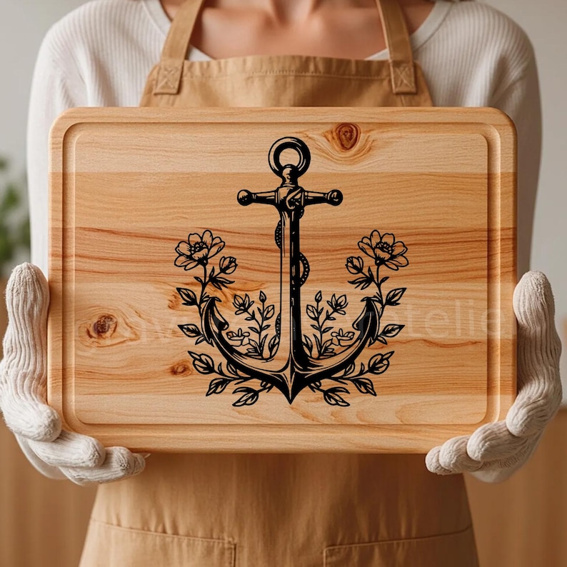 Puede incluir: Una tabla de cortar de madera con un dise&ntilde;o de ancla negra rodeada de elementos florales. El ancla tiene un detalle de cuerda y las flores son estilizadas.