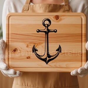 Puede incluir: Una tabla de cortar de madera con un dise&ntilde;o de ancla negra. La tabla tiene una veta de madera natural y un borde redondeado.