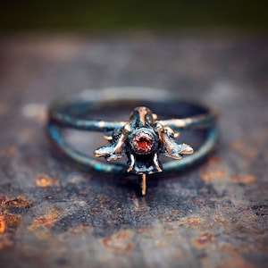 May include: A ring made of metal with a dark blue patina, featuring a detailed anatomical design of a vertebra with a red gemstone in the centre.